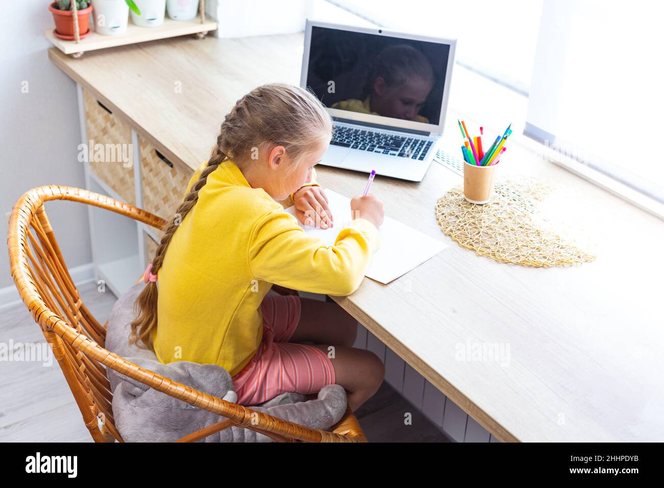 I bambini scrivono nel notebook, seduti alla scrivania accanto alla finestra. La ragazza della scuola caucasica in felpa gialla sta facendo i compiti a casa di fronte al laptop. Casa Foto Stock