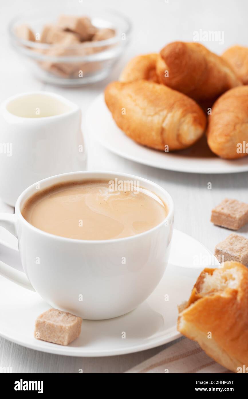 Caffè con latte in una tazza bianca, panini e zucchero di canna su un tavolo di legno bianco. Foto Stock