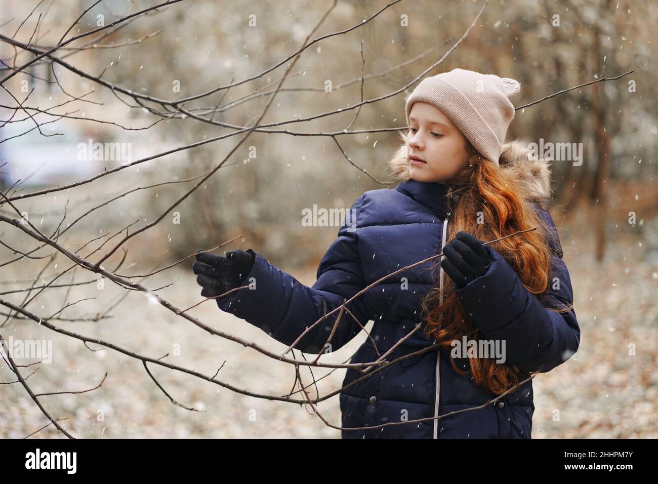 Ragazza piccola carina che tiene un ramo di albero senza foglie su uno sfondo di fiocchi di neve caduta. Primo concetto di neve. Foto Stock