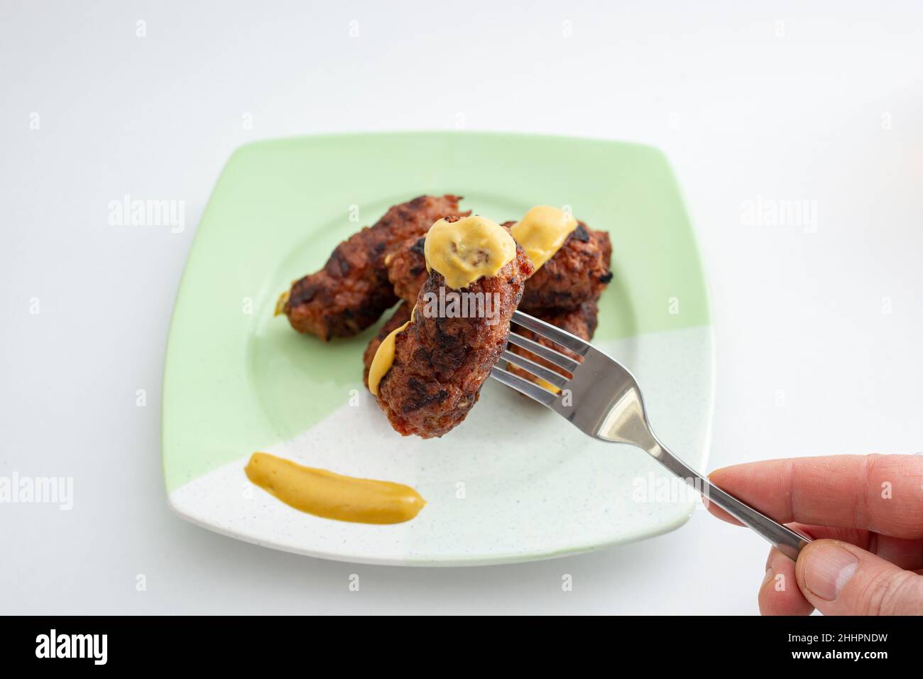 Mici o mitititititei (cibo tradizionale rumeno) con senape su un piatto di ceramica, su sfondo bianco. Tenere a mano una forcella in acciaio inox Foto Stock