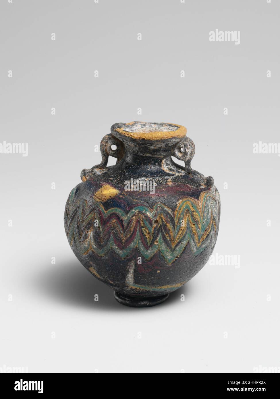 Vetro Aryballos (bottiglia di profumo) 5th secolo a.C. Greco, Mediterraneo orientale viola scuro opaco, appare nero, con striature di colore rosso marrone più chiaro, con le stesse maniglie di colore; sentieri in giallo opaco e blu turchese opaco. Disco-bordo inclinato verso l'interno con bordo sporgente verso la bocca ruvido; collo concavo corto; spalla larga inclinata; corpo quasi sferico; convesso, Fondo leggermente disuniforme; due manici ad anello con code con nodi si estendono dalla spalla al collo e dalla parte inferiore del cerchione-disco.una traccia gialla applicata al bordo esterno del cerchione-disco, con la parte superiore del collo ripiegata su una parte della traccia; traccia gialla e blu turchese Foto Stock