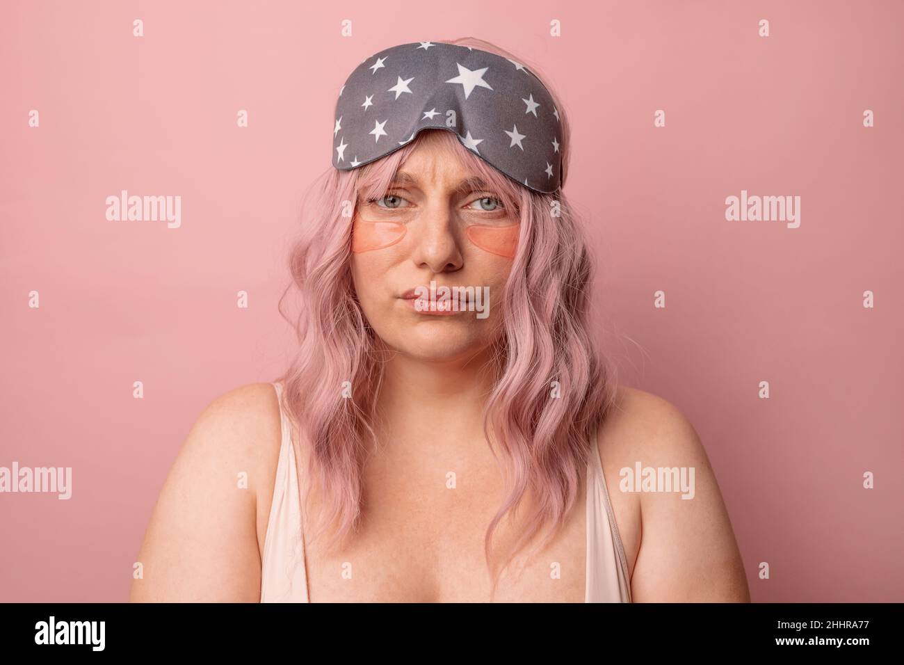 Sonnolenta addormentata femmina caucasica indossa la maschera del sonno, triste svegliarsi presto al mattino posa sopra rosa studio parete sfondo. Foto Stock