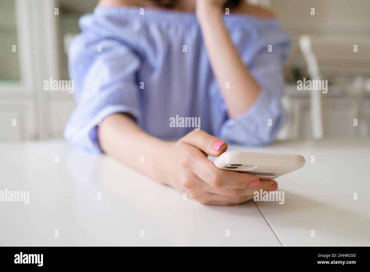 Giovane donna in elegante blusa blu con manicure pulito tiene smartphone moderno seduto al tavolo bianco al mattino in piano camera Foto Stock
