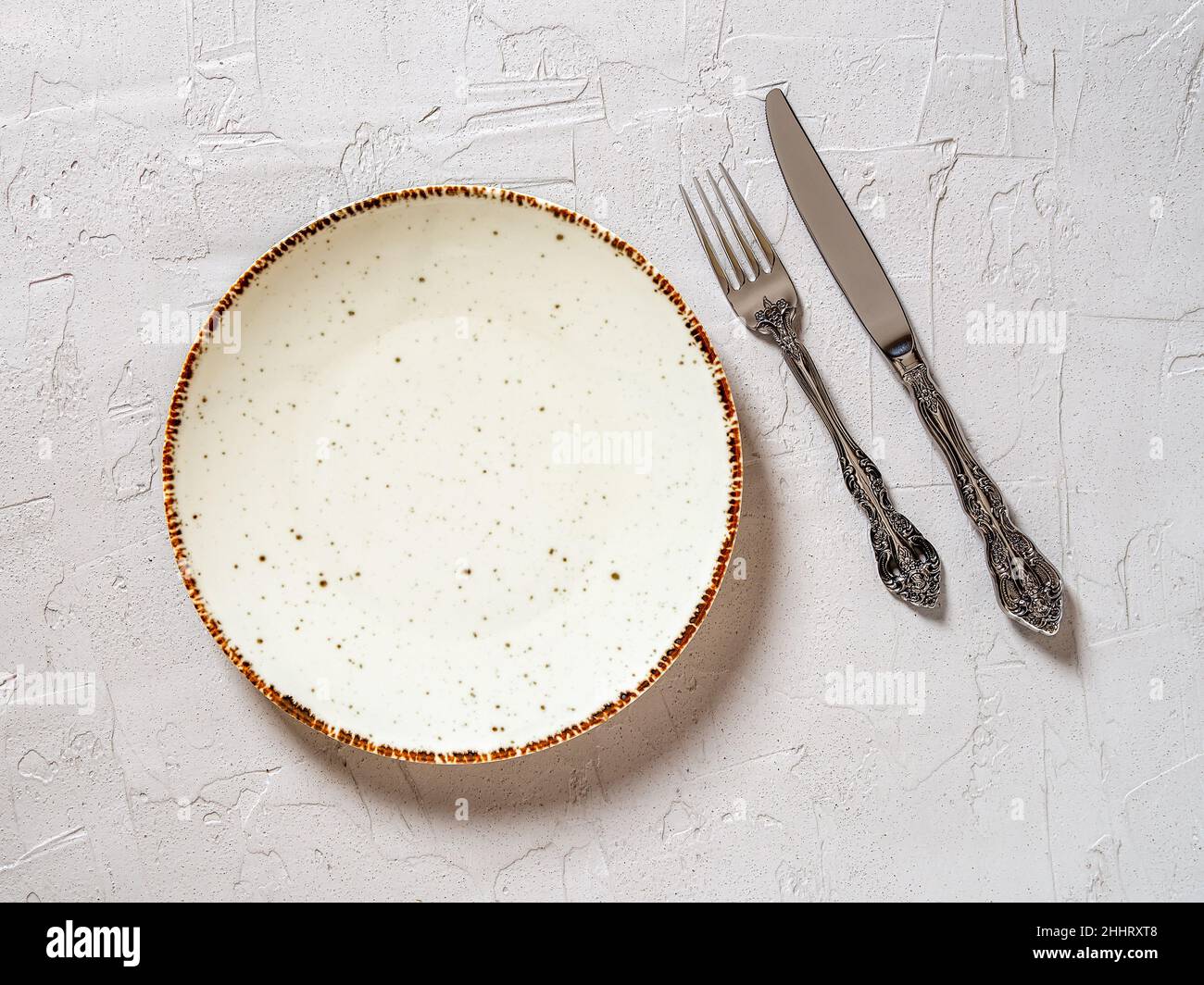 Piatto da pranzo in porcellana bianca, coltello da tavolo vintage e forchetta su sfondo grigio testurizzato. Svuotare la piastra puntinata vicino a un coltello elegante e a una forcella. Vista dall'alto. Foto Stock