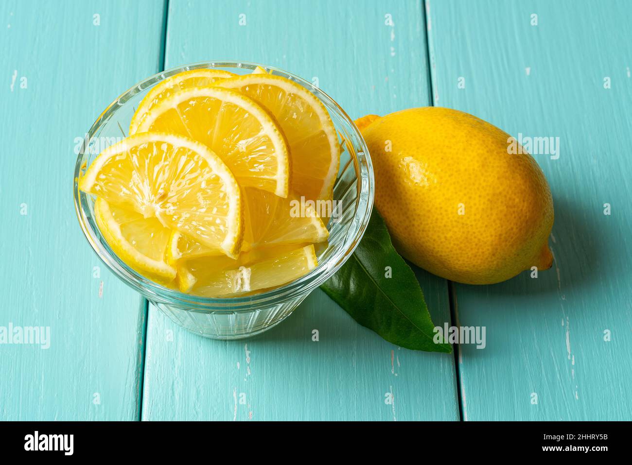 Fette di limone crude in un recipiente di vetro e una frutta intera su un tavolo di legno turchese. Agrumi freschi di colore giallo tagliato per concetti di vitamina C, antiossidante. Foto Stock