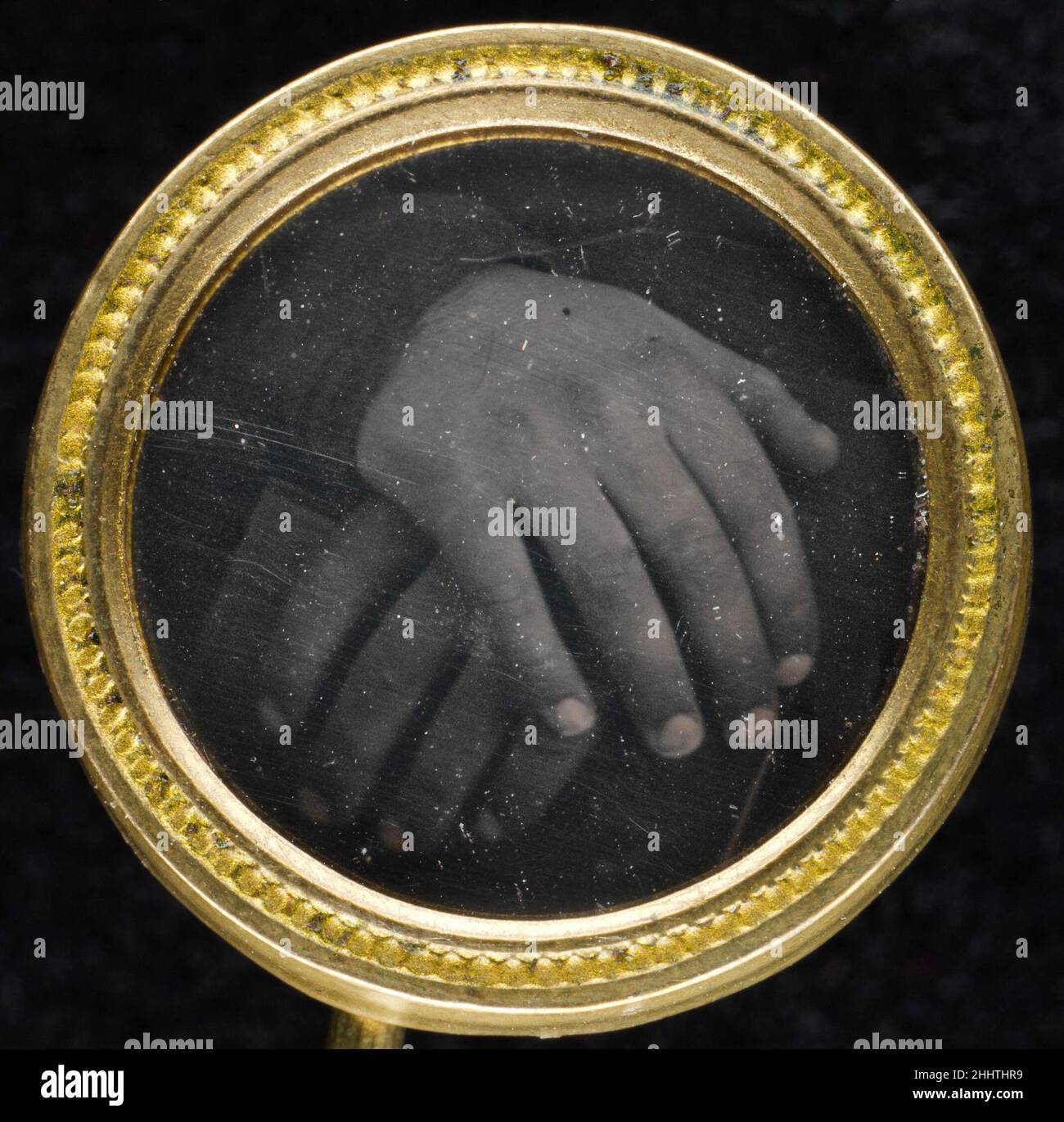 [Button] 1840s–50s Sconosciuto questo daguerreotipo in miniatura mostra due mani poggiate su un libro. La fotografia è impostata in una struttura in ottone a due pezzi lavata in oro con un anello sul retro per cucire ad un indumento. Il design del contenitore con il suo bordo ornamentale semplice e rialzato è tipico dei bottoni in metallo dorato prodotti in serie dal 1830 al 1850 in diverse fabbriche del New England come la Scovill Manufacturing Company di Waterbury, Connecticut, che fabbricava anche lastre di daguerreotype. Il pulsante è stato scoperto all'inizio del 1980s in un mercato delle pulci in Massachusetts. L'uso di pulsanti per i purpos politici Foto Stock