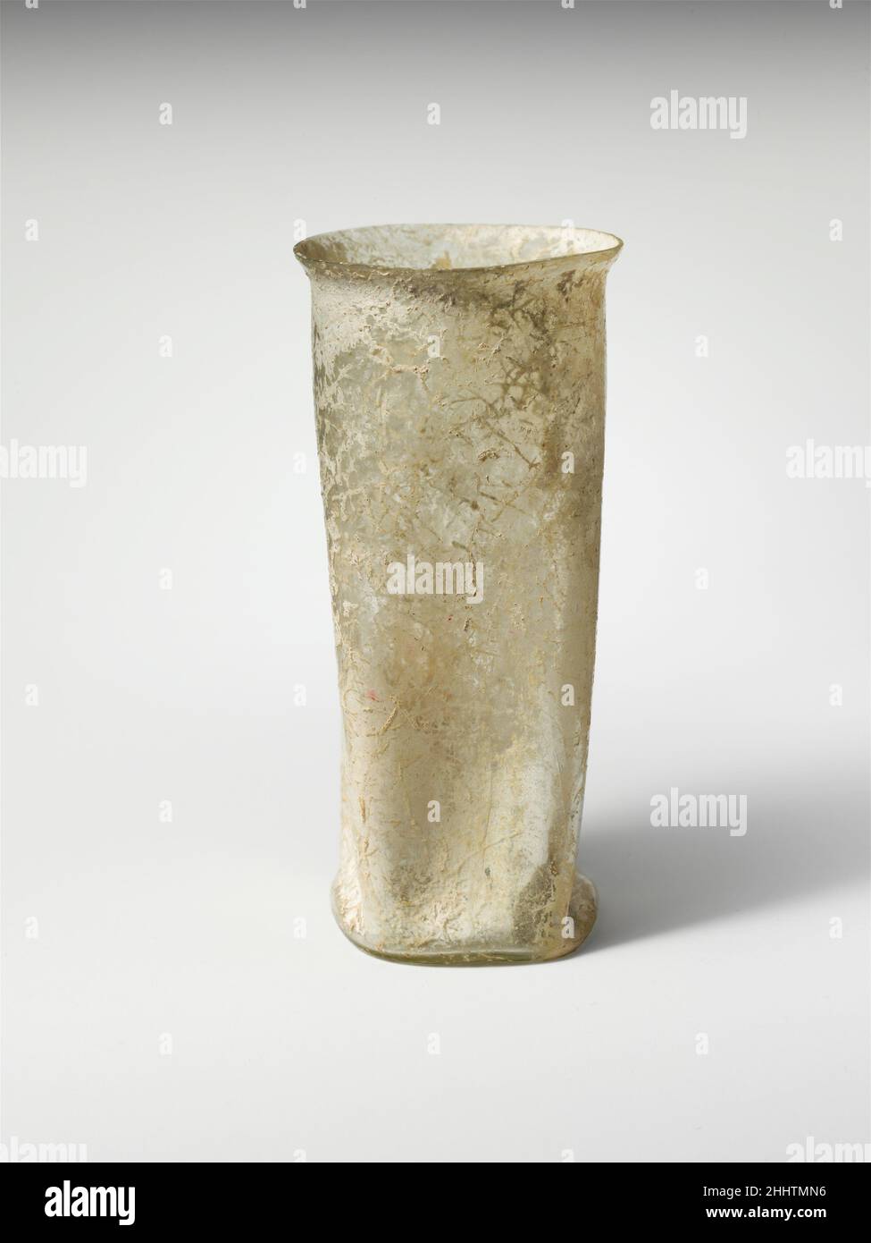 Bicchiere di vetro 2nd–3rd secolo d.C. romano incolore con tinta verde pallido.bordo spaccato, incrinato e irregolare; lato verticale, cilindrico in alto, quindi sagomato in un quadrato grezzo con angoli cavi; fondo quadrato, spinto in rilievo, bordo arrotondato e piccolo segno di pontil al centro.sotto il bordo, due fasce orizzontali di linee abrase ruote; Sul corpo, quattro rientranze lunghe, formando una forma a stella-come ad interior.intact, tranne per piccolo chip di intemperie nel bordo e crepa nel corpo inferiore; pinprick e bolle più grandi; incrostazione di suolo e segni di radice che coprono la maggior parte delle superfici, e iridence debole.. Vetro bea Foto Stock