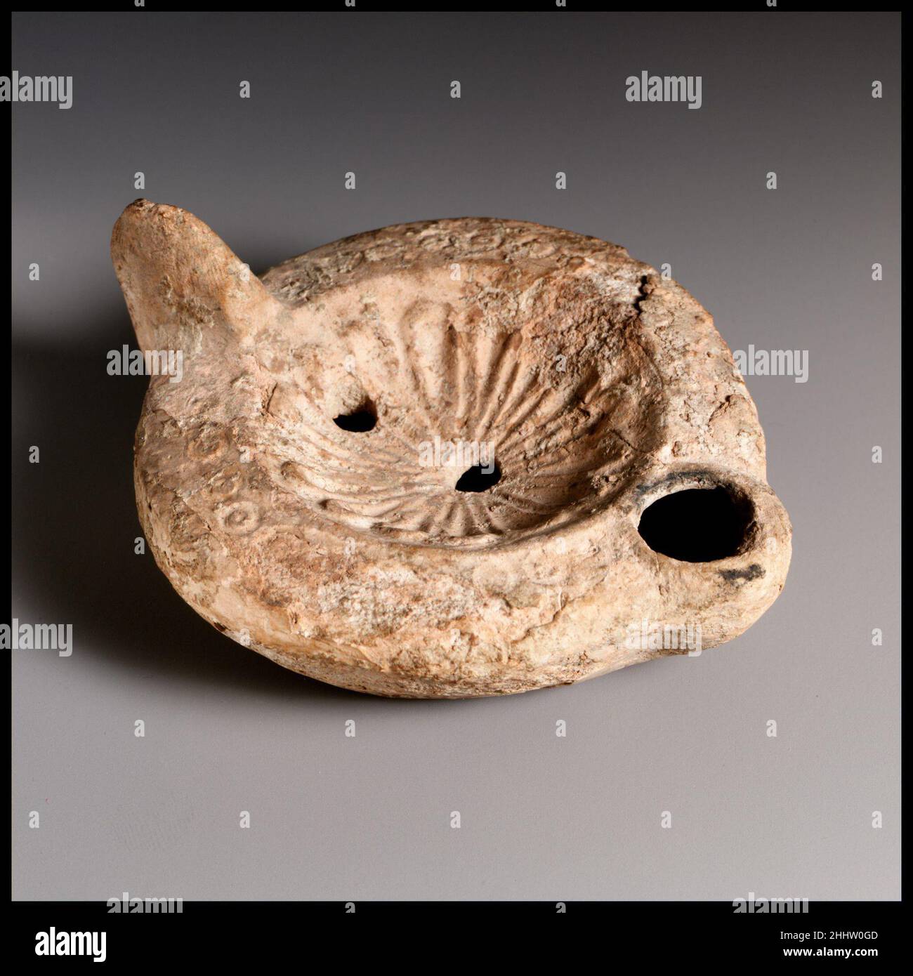 Lampada ad olio di terracotta fine 3rd secolo d.C. Roman Loeschcke tipo 8. Impugnatura non forata. Realizzato in stampo. Disco; rosetta con petali cavi e irradianti attorno al foro centrale di riempimento; un secondo foro di riempimento verso il bordo davanti al manico. Sulla spalla: Una fila di cerchi e punti impressi. Su una base concava irregolare all'interno dell'anello di base scritto in greco: ]OM[o], con linee sopra e sotto. Grave accumulo e vaiolatura di superfici. Lampada ad olio di terracotta. Romano. Fine 3rd secolo d.C.. Terracotta. Tardo imperiale. Terracotte Foto Stock