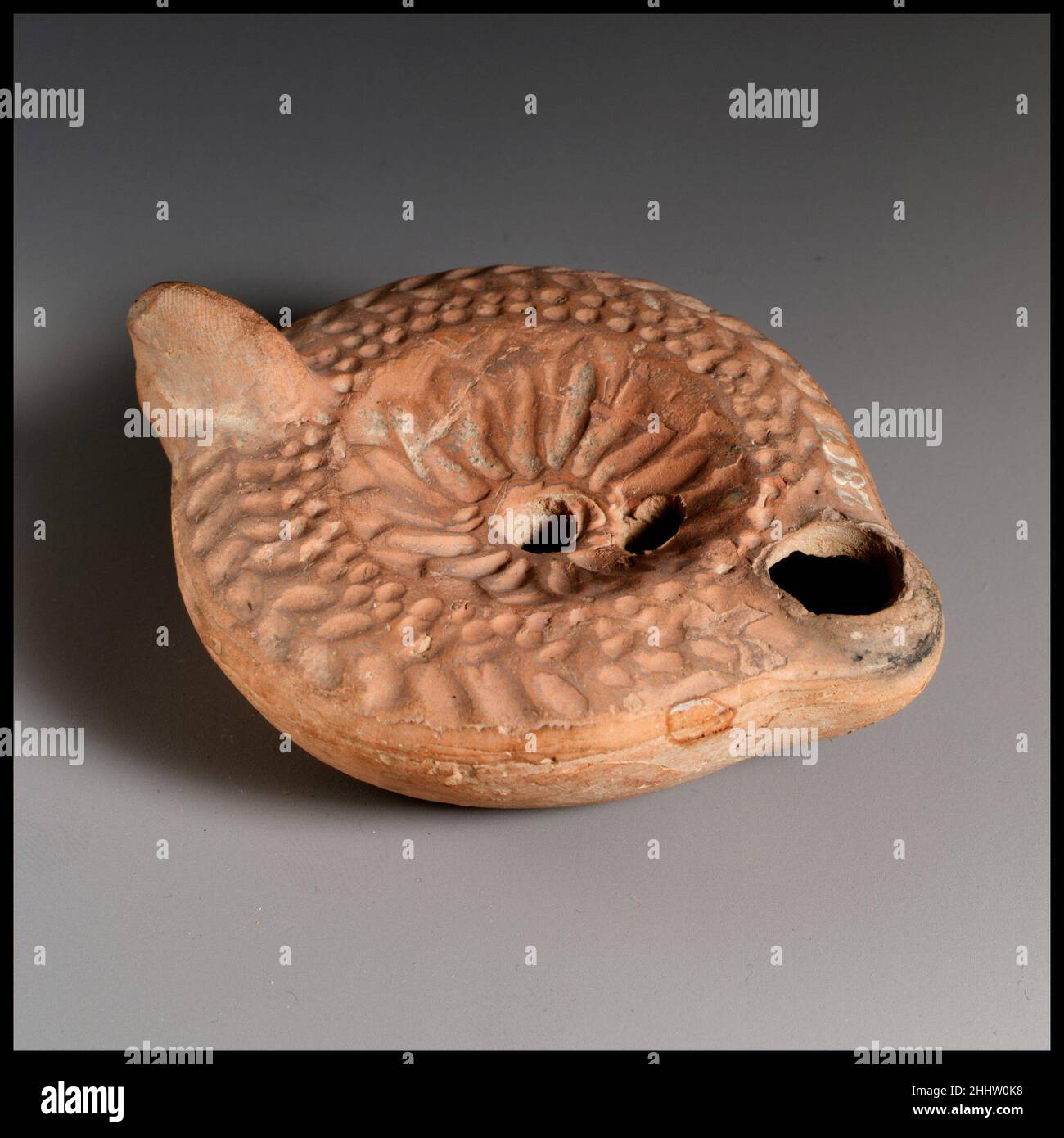 Lampada ad olio di terracotta fine 3rd secolo d.C. romana, Cypriot Loeschcke tipo 8. Impugnatura non forata. Realizzato in stampo. Disco; rosetta con petali cavi e irradianti attorno al foro centrale di riempimento; un secondo foro di riempimento verso l'ugello. Sulla spalla: Due file di puntini impressi circondate da una corona di aringhe. Base concava profonda inscritta crudelmente in lettere greche: C?YXI/?WNOC.. Lampada ad olio di terracotta. Romano, cipriota. Fine 3rd secolo d.C.. Terracotta. Tardo imperiale. Terracotte Foto Stock