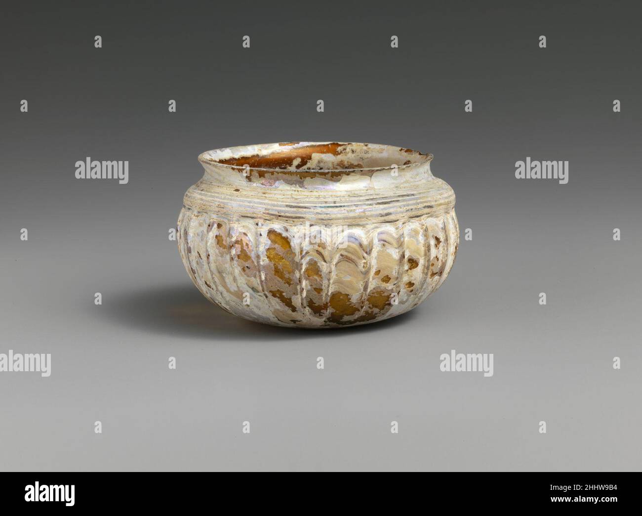 Coppa con nervature di vetro metà 1st secolo d.C. romana traslucido giallo miele; sentiero in bianco opaco.Knocked-off, bordo smerigliato; collo concavo corto; collare a rigonfiamento orizzontale sotto; corpo globulare squat curvato verso fondo quasi piatto.Sentiero avvolto a spirale dal basso intorno al corpo al collo, spesso in fondo, ma sottile e sottile in alto; ventisette, poco profonde, leggermente irregolari, nervature verticali, Che si estende dal rigonfiamento sotto il collo alla sottobesa di side.intact, ad eccezione di un piccolo chip di intemperie nel bordo; alcune bolle; dulling e vaiolatura di bolle di superficie, la maggior parte delle superfici ricoperte di intemperie bianche spesse e ir Foto Stock