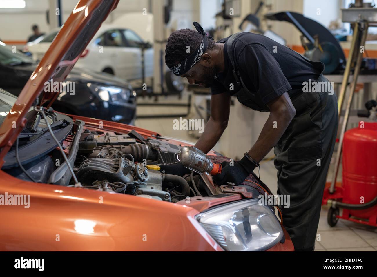 Giovane uomo africano in abbigliamento da lavoro che si piega sul cofano dell'auto aperto durante il controllo del motore o la riparazione dei dettagli del veicolo Foto Stock