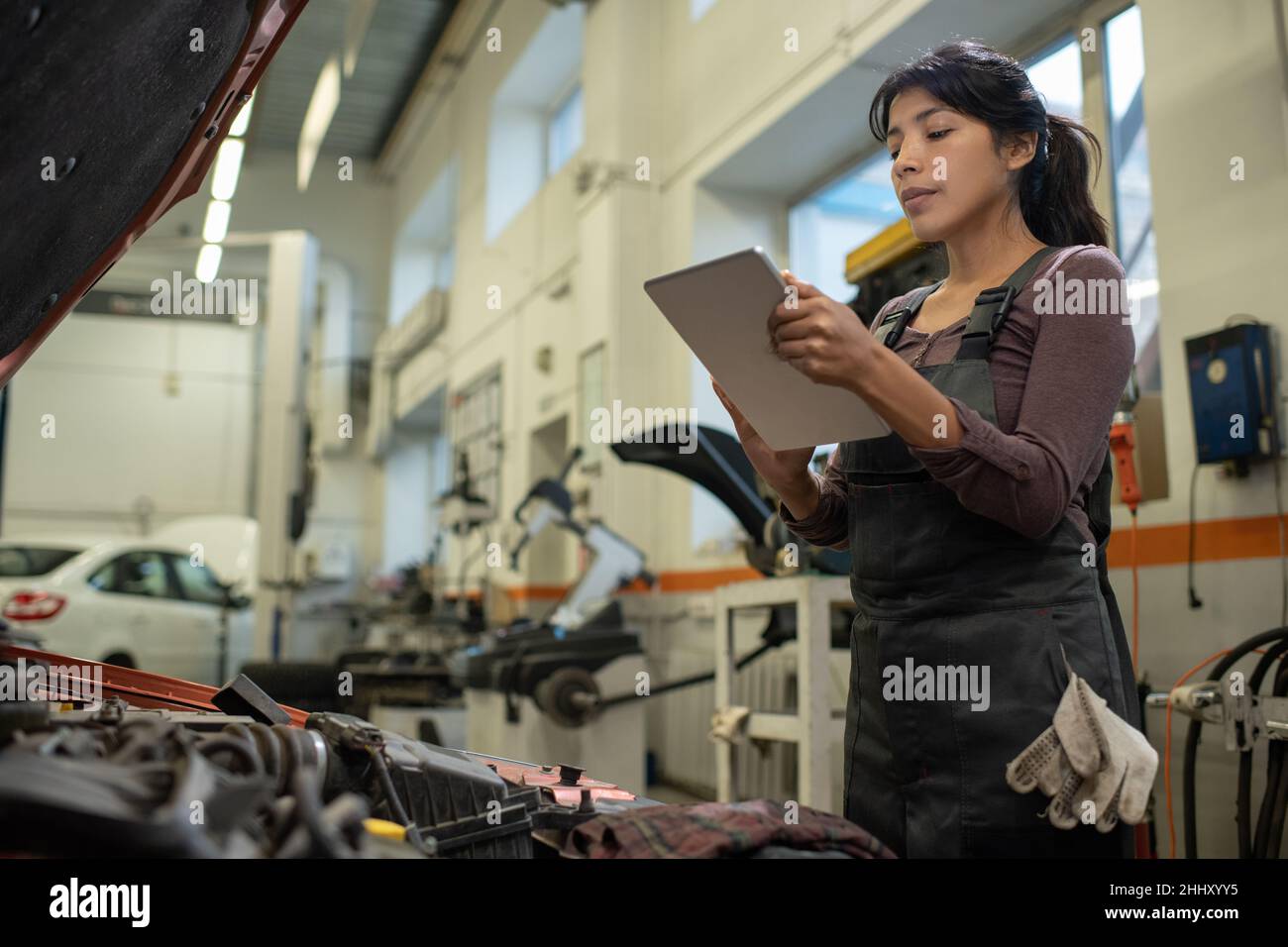 Giovane ispanica donna in tute che guarda lo schermo di tablet digitale nelle sue mani mentre ripara il motore rotto dell'auto Foto Stock