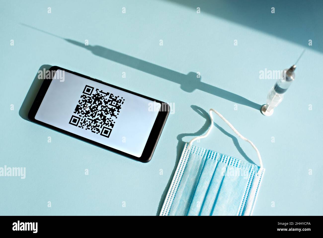 Smartphone con codice quar di certificato di vaccinazione sullo schermo, siringa e maschera medica su sfondo blu chiaro. Foto Stock
