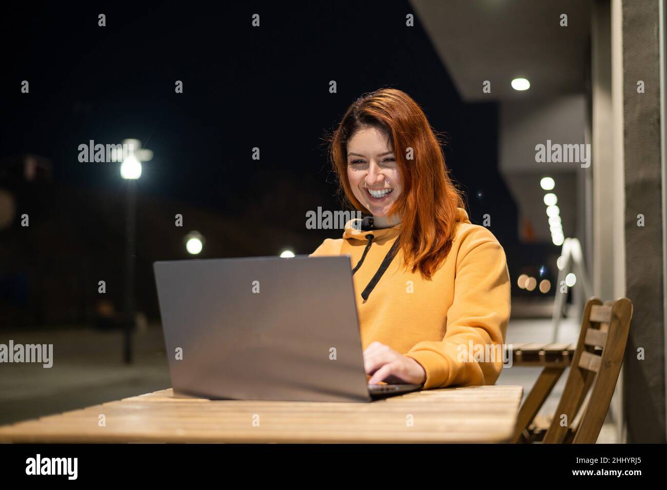 Una donna lavora a distanza a un computer portatile in un bar estivo a tarda sera. Ragazza felice studiare mentre si siede su una strada vuota ad un tavolo di legno Foto Stock