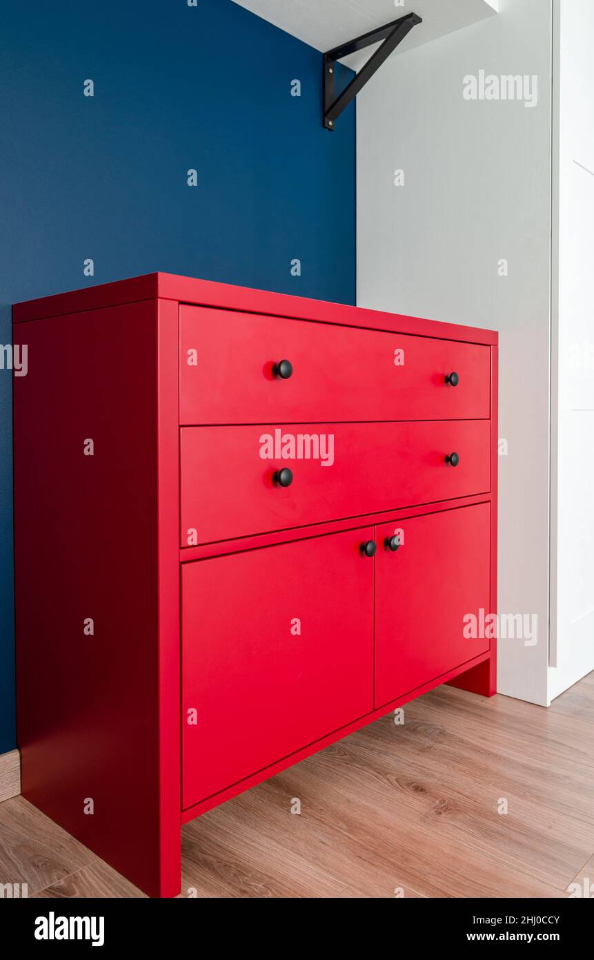 Cassettiera rossa in un interno scandinavo. Un armadietto di legno rosso  con le maniglie nere si leva in un appartamento contro una parete blu  vicino ad una wa bianca Foto stock -