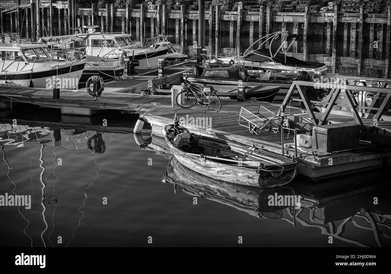 Un dinghy usurato è ormeggiato ad un molo e si riflette nell'acqua still. C'è una bicicletta sul molo e le barche oltre. Foto Stock