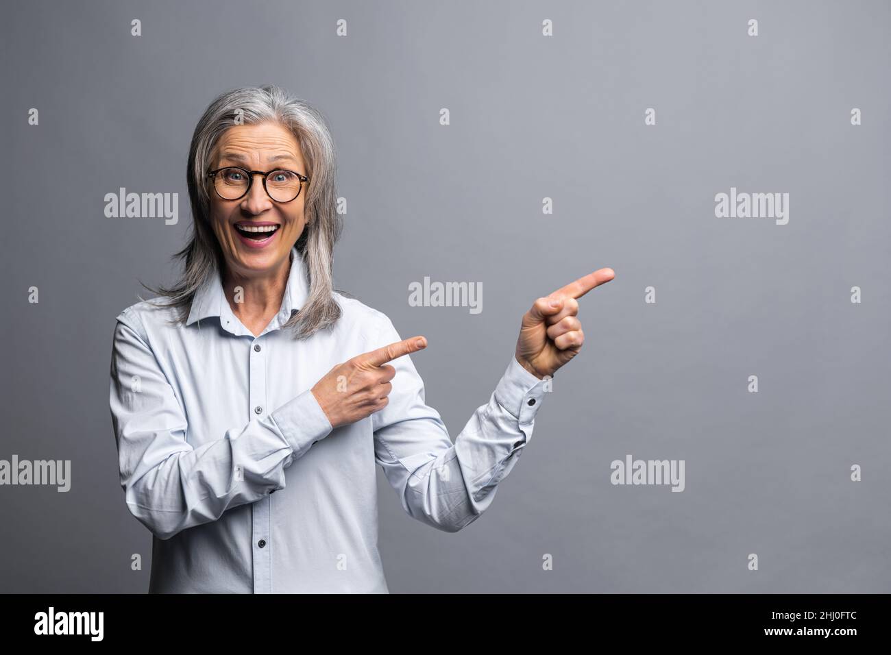 Donna d'affari positiva indossando abiti di stile ufficiale puntando dito lontano e prestando la vostra attenzione a spazio vuoto per la pubblicità mentre si guarda la macchina fotografica. Studio girato isolato su grigio Foto Stock
