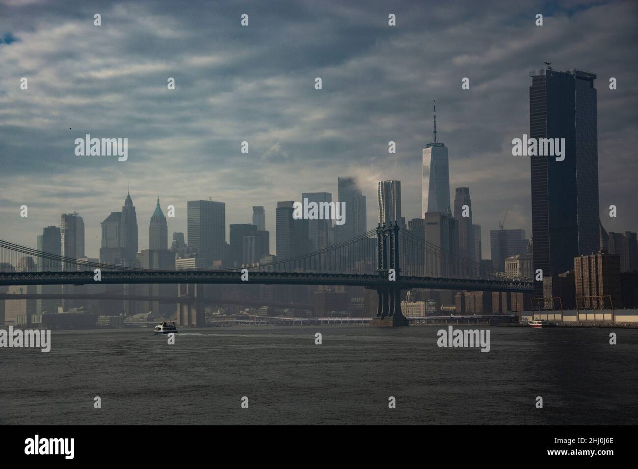 Dramatischer Blick von der Fähre auf dem East River auf die beiden Stahlbrücken und Lower Manhattan und dem imposanten One World Trade Center Foto Stock