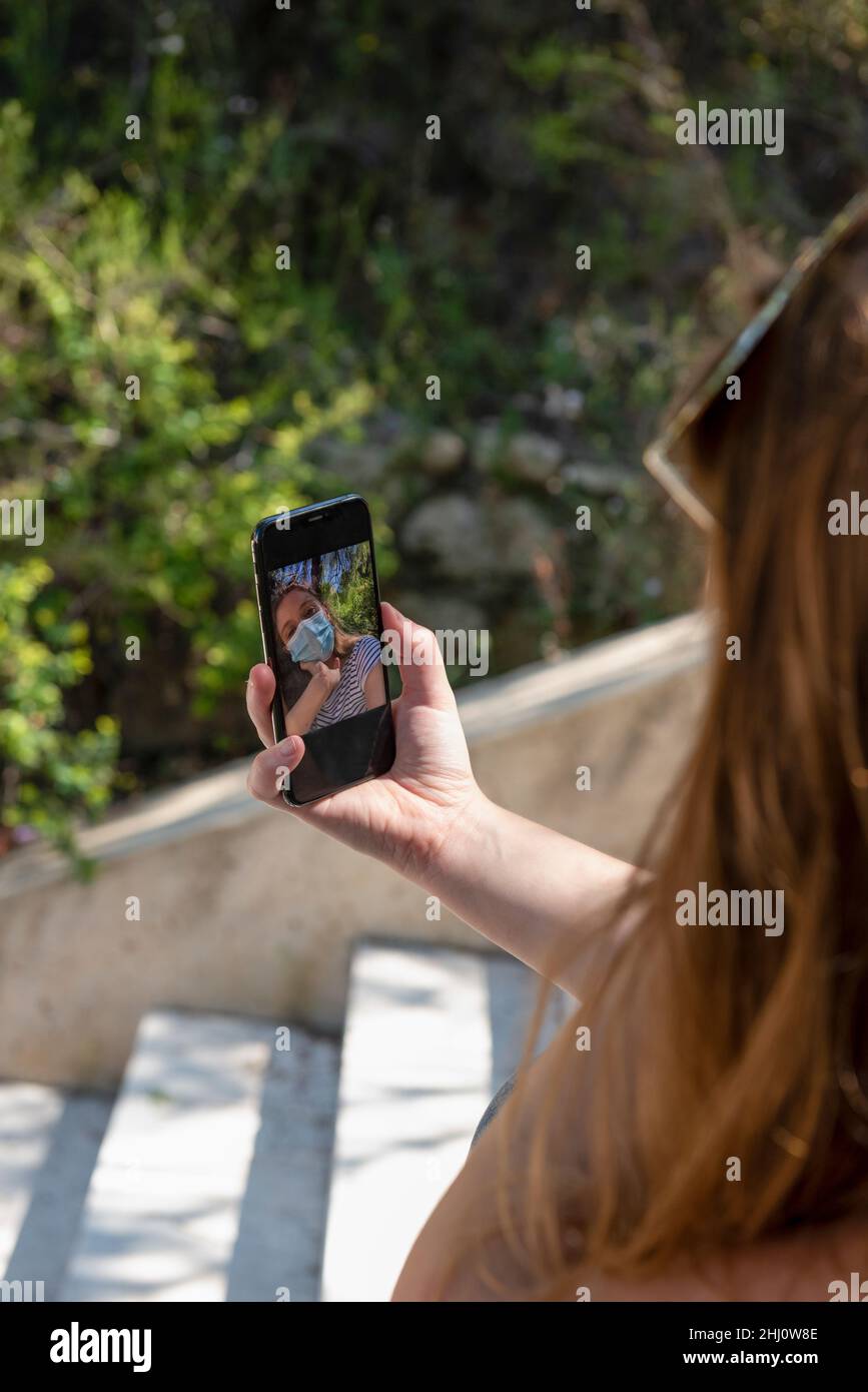 Donna irriconoscibile con maschera facciale utilizza lo smartphone per prendere selfie. Foto Stock