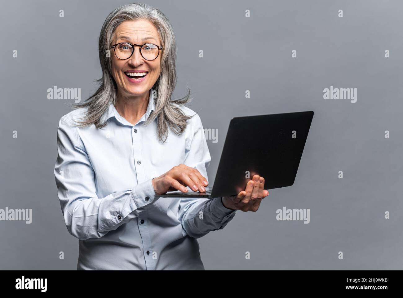 Donna anziana moderna in abbigliamento formale utilizzando computer portatile isolato in grigio. Ritratto di una donna matura dipendente ufficio utilizzando la tecnologia online per fare affari, computer app per la contabilità Foto Stock