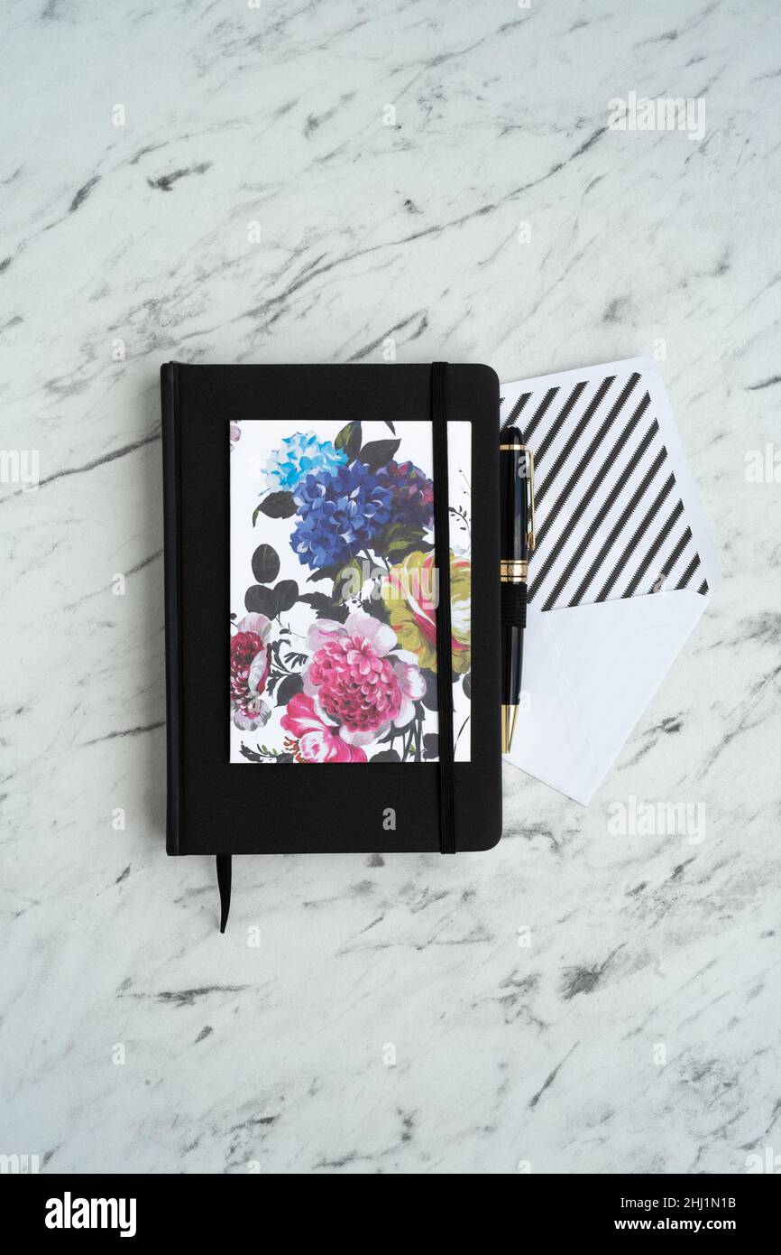 Blocco note nero al centro del tavolo in marmo, accanto ad una carta luminosa con fiori, una busta bianca e una penna elegante Foto Stock