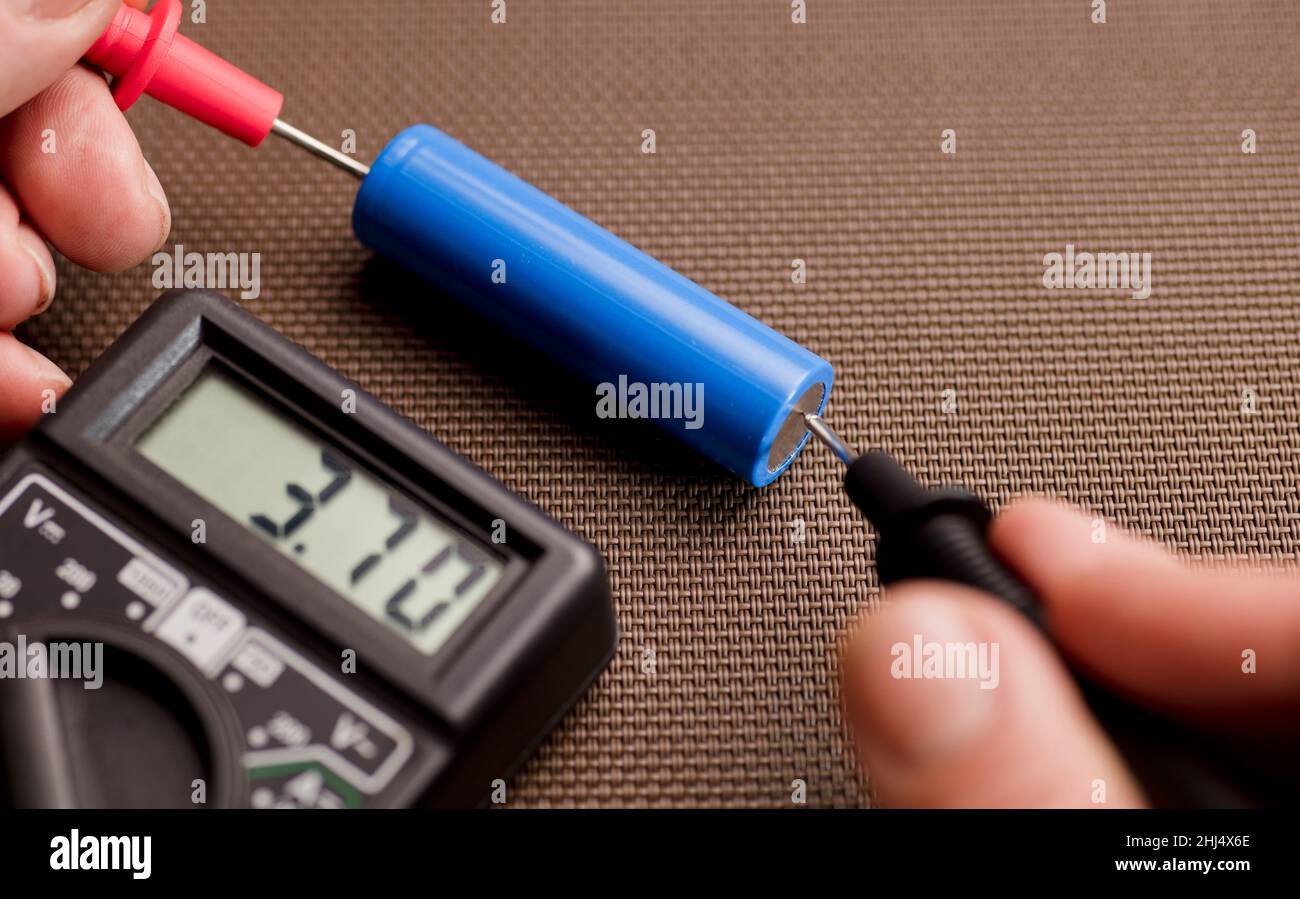 batteria con calcolatore da volt, voltmetro nelle mani di un uomo, polo positivo rosso, polo negativo nero, su un tavolo da lavoro Foto Stock