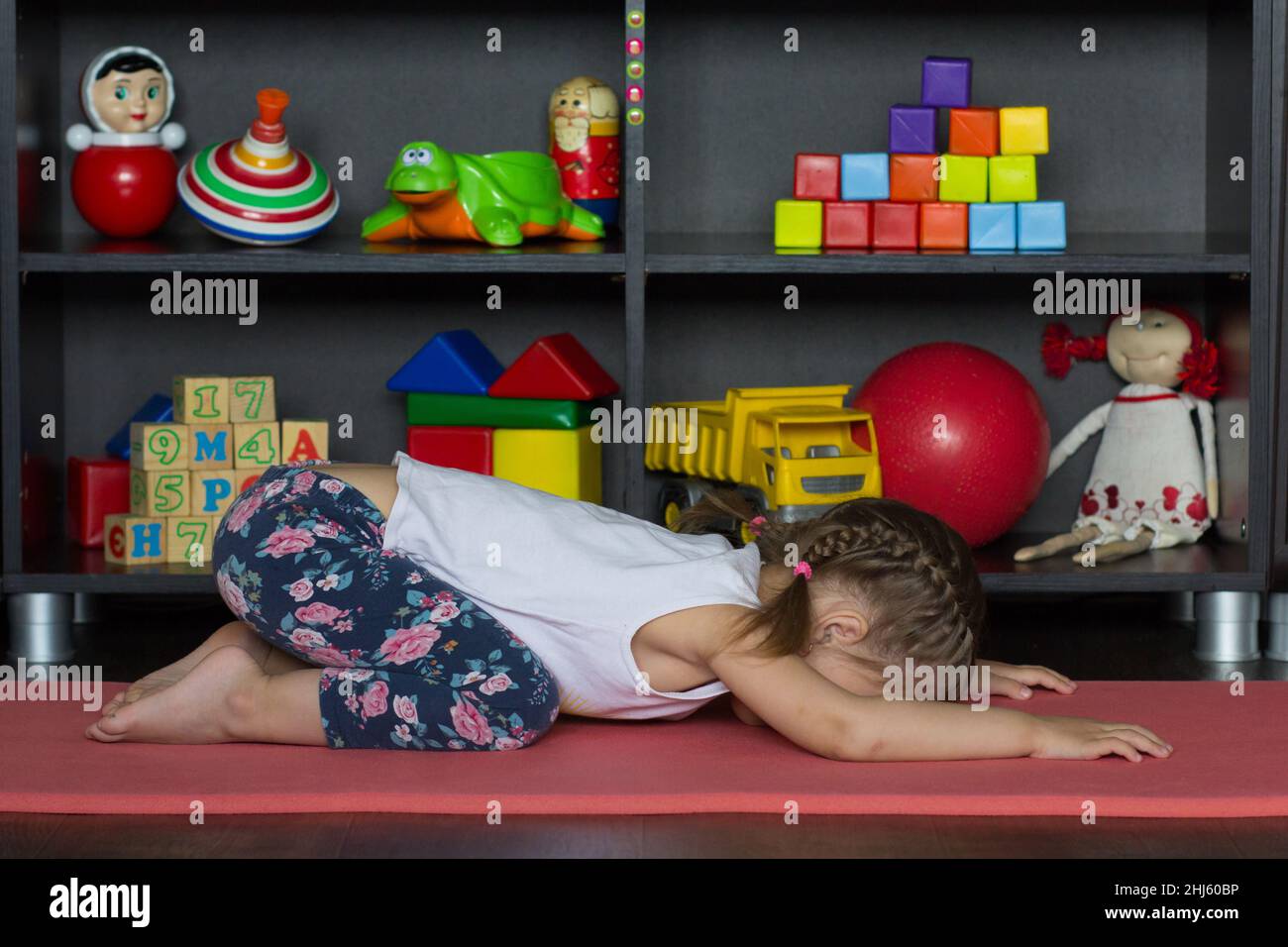 La bambina si trova in posa estesa di yoga bambino come sedersi su tacchi, portare la fronte giù per riposare di fronte alle ginocchia, posizionare palmo di mani piatto davanti a h Foto Stock