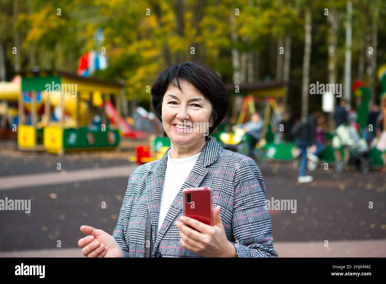 Ritratto di una senora felice per 50 anni nel parco in autunno in una giacca grigia sul parco giochi. Una donna ride e usa uno smartphone rosso Foto Stock