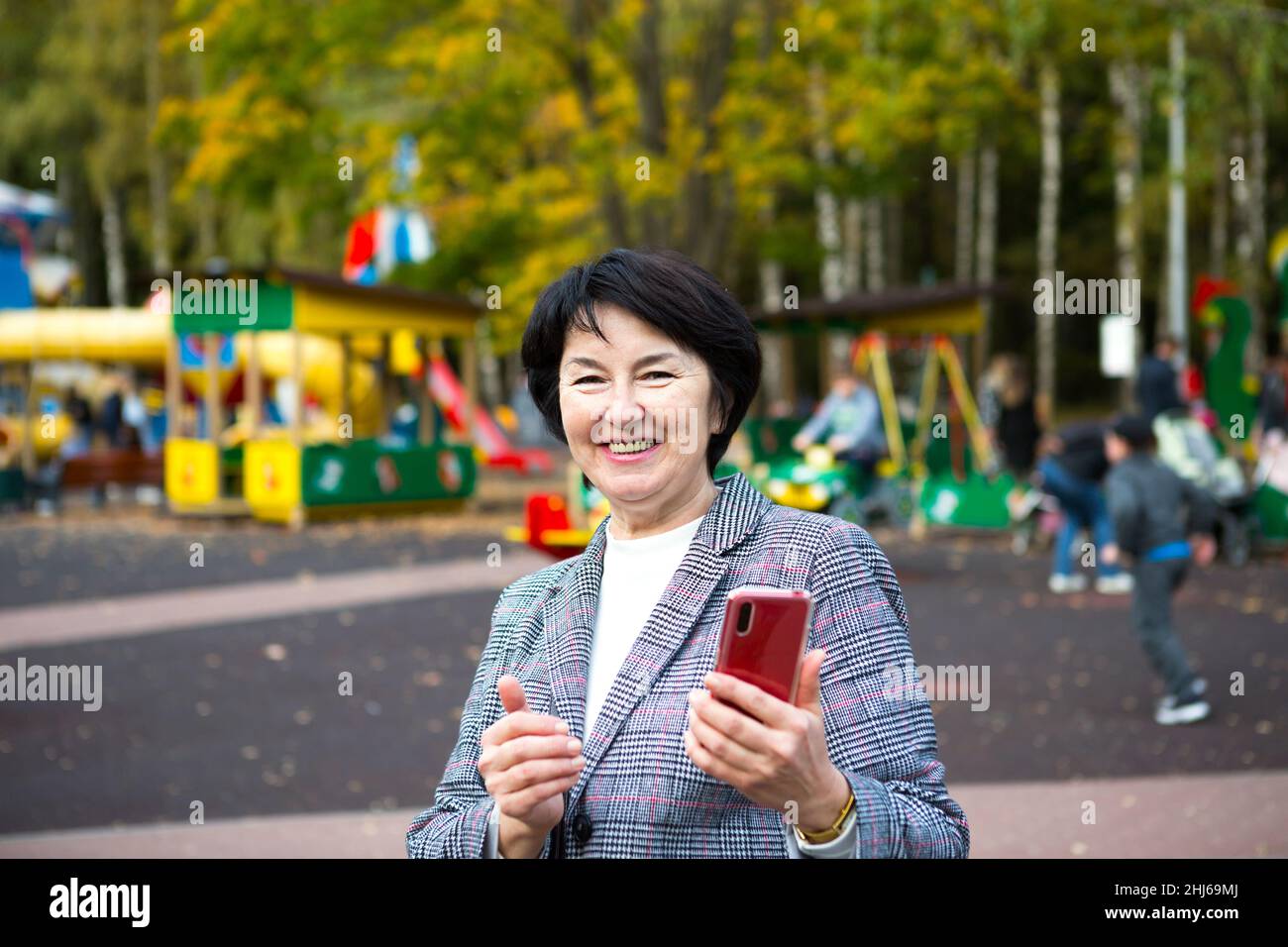 Ritratto di una senora felice per 50 anni nel parco in autunno in una giacca grigia sul parco giochi. Una donna ride e usa uno smartphone rosso Foto Stock