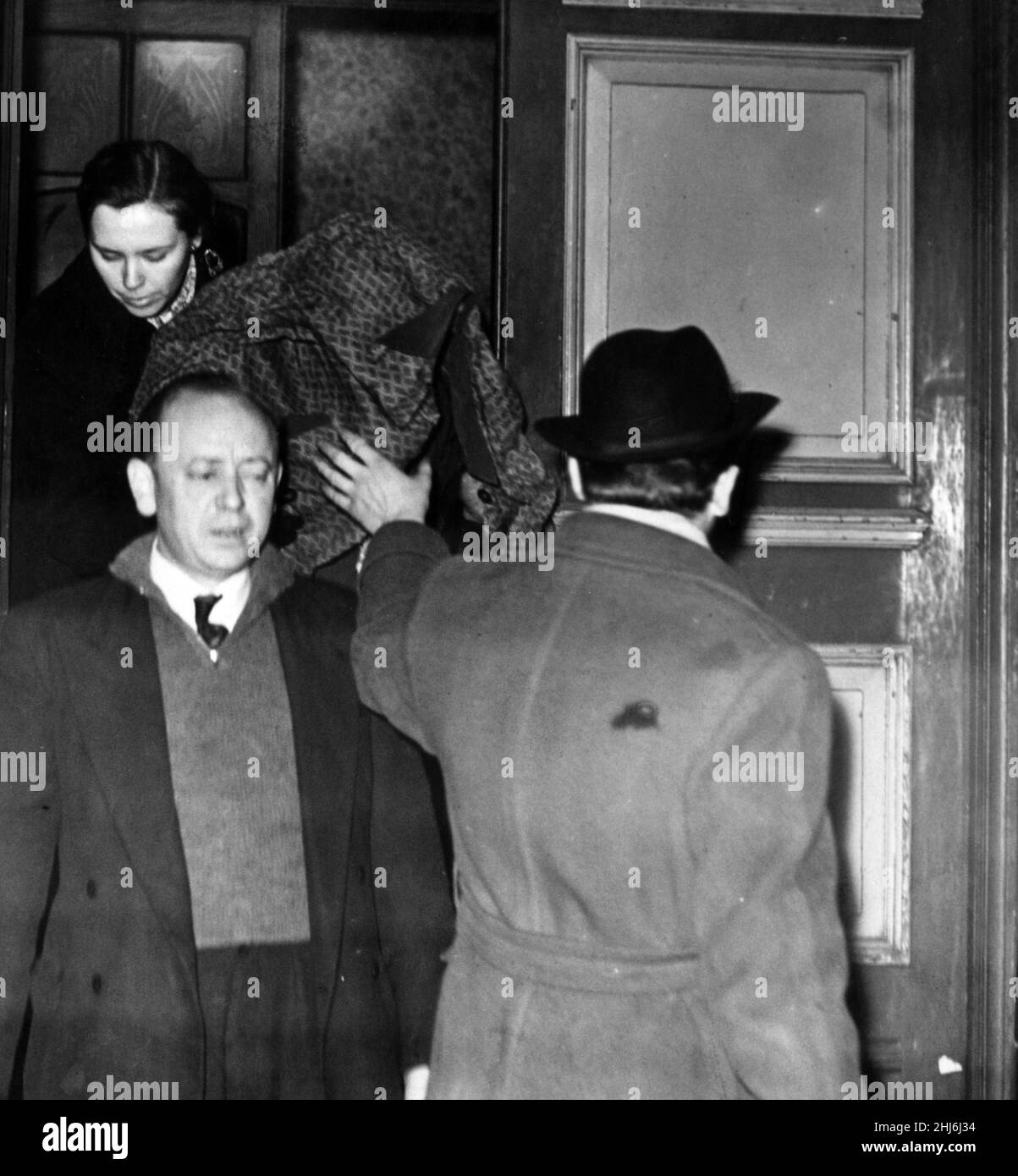 Due poliziotti scortano la signora Wilson, la sua testa coperta da un impermeabile, dall'hotel ad un'auto d'attesa. 12th dicembre 1957. Foto Stock