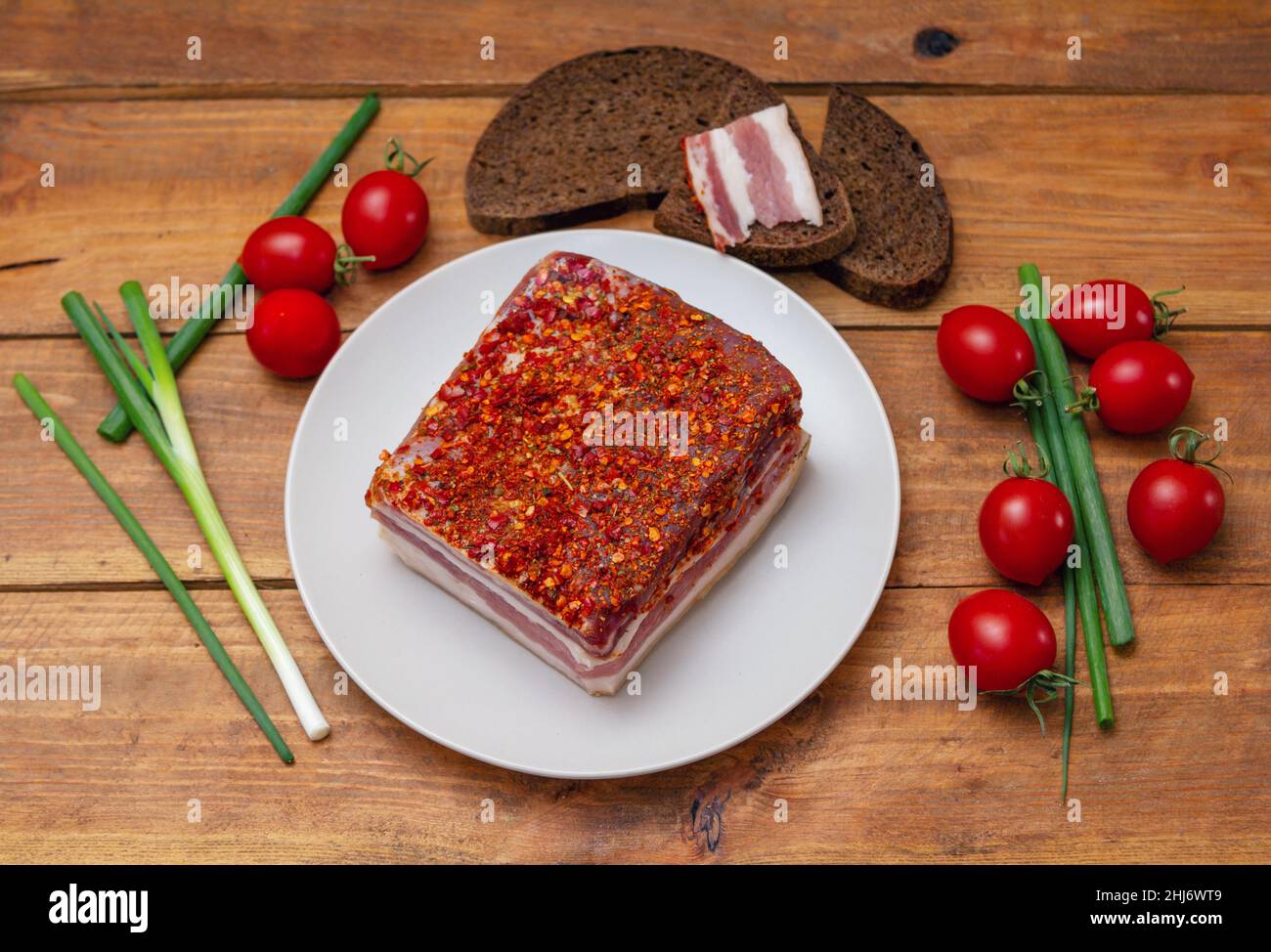 Pancetta piccante affumicata su un piatto. Fetta di carne di maiale affumicata su pane, pomodori ciliegini e cipolla verde su tavola di legno. Foto Stock