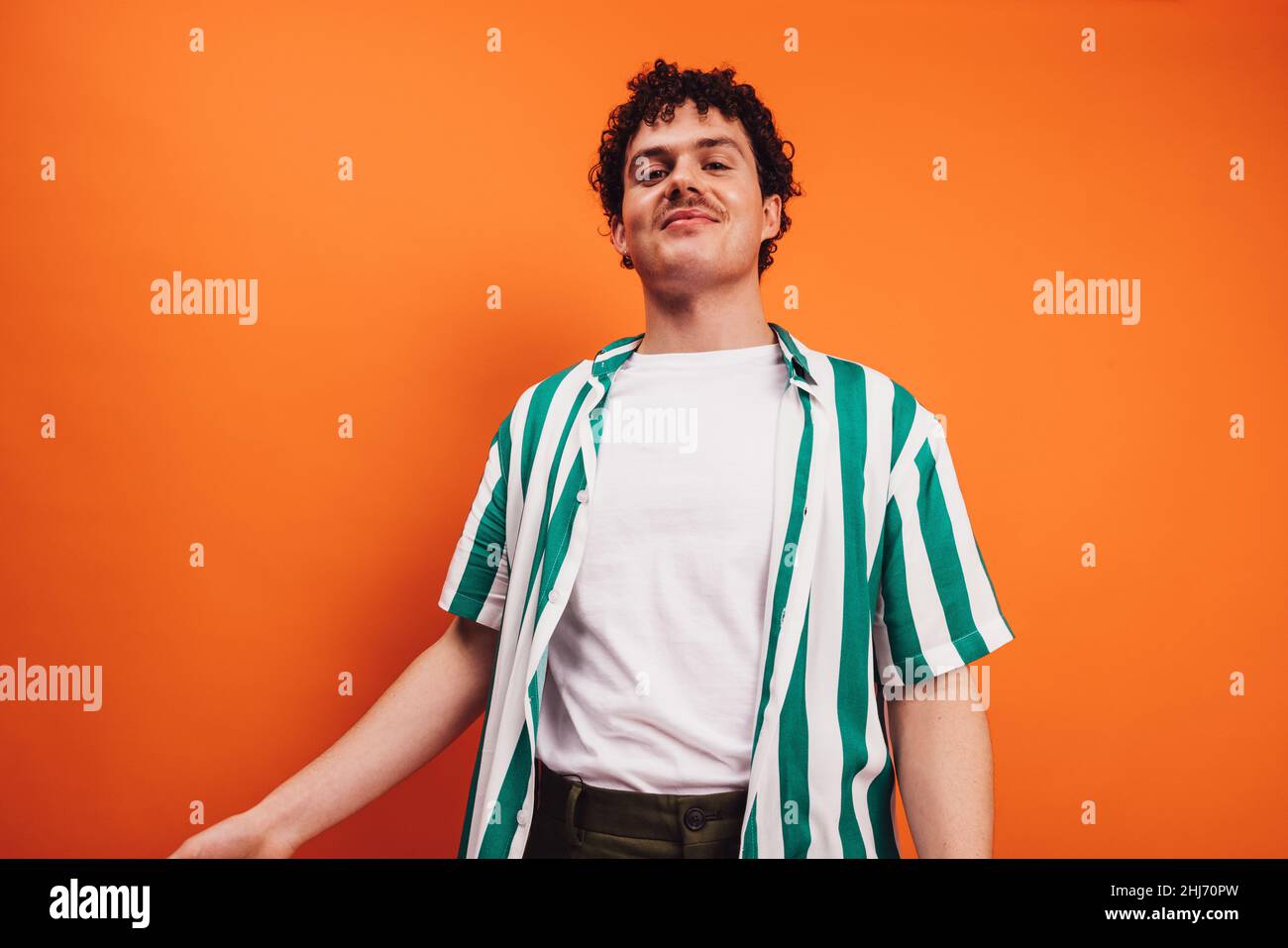 Giovane queer che guarda la fotocamera con un volto allegro in uno studio. Felice germogli della giovane generazione z sorridente mentre si levano in piedi da solo contro un orang Foto Stock