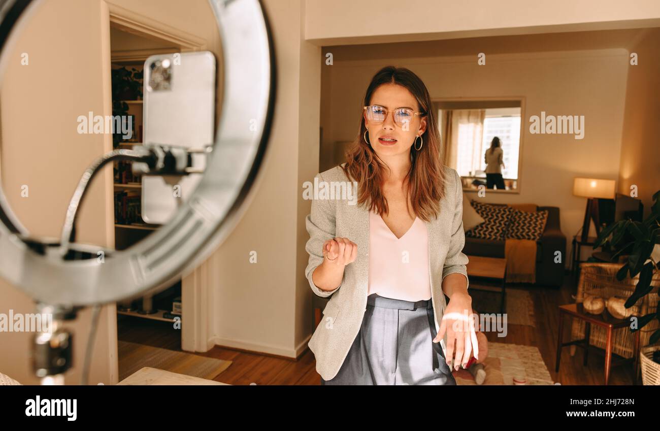 La mamma millenaria registra contenuti per il suo canale di social media. Influencer femminile che parla di fronte a una lampada ad anello con il bambino che gioca nel backgro Foto Stock