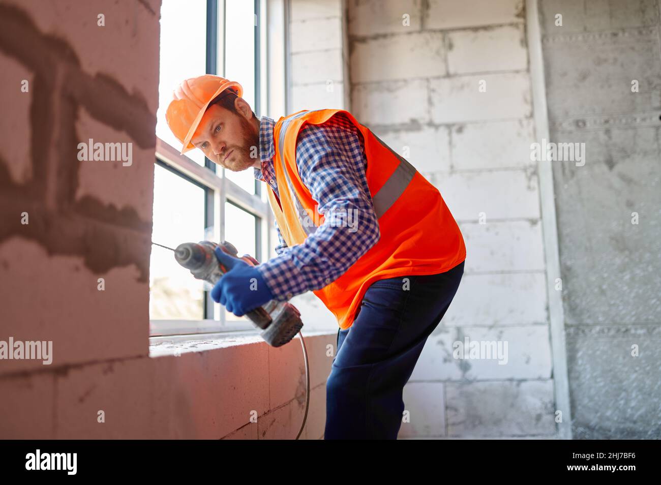 Costruttore in hardhat che lavora con il trapano in ambienti chiusi Foto Stock