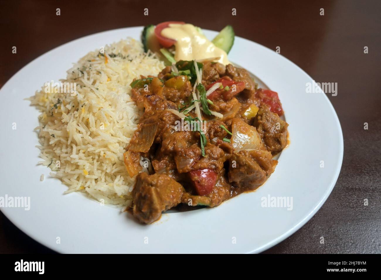 Curry indiano a base di carne e verdure in salsa piccante servito con decorazione di riso e insalata su un piatto bianco e un tavolo di legno marrone, selezionato Foto Stock