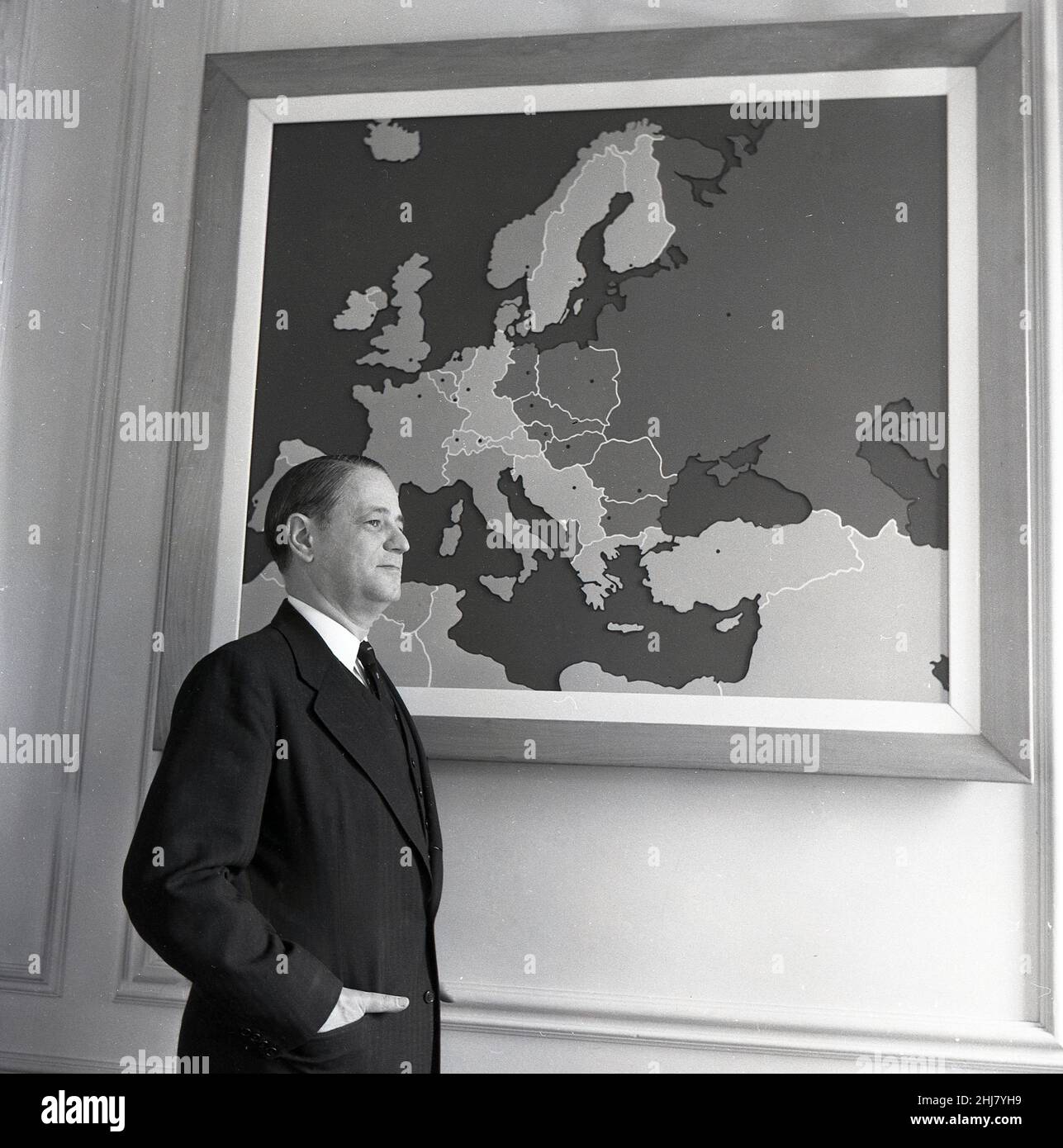 1960s, storico, all'interno di una stanza, un gentleman in tuta in tre pezzi, l'Ambasciatore degli Stati Uniti alla NATO in piedi da un quadro incorniciato sul muro che mostra una mappa dell'Europa. Foto Stock