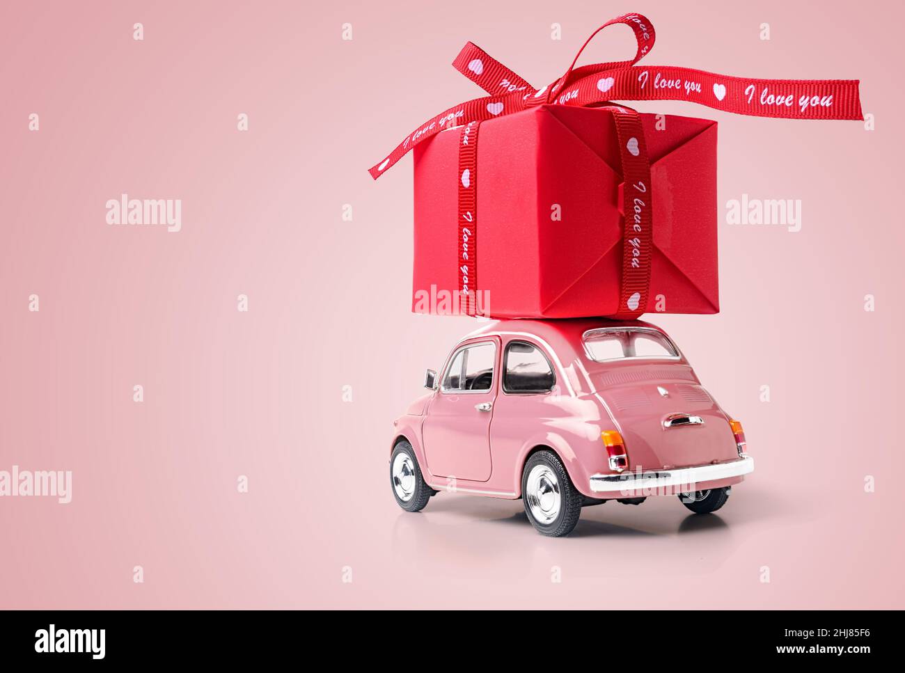 Macchina giocattolo retro rosa che consegna scatola regalo rossa con nastro su sfondo rosa. Spazio di copia. Foto Stock