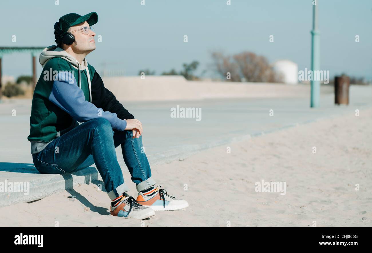 un uomo di mezza età, che indossa una felpa con cappuccio e un paio di jeans multicolore, ascolta la musica utilizzando un paio di cuffie seduti sul lungomare, accanto alla sabbia di Foto Stock