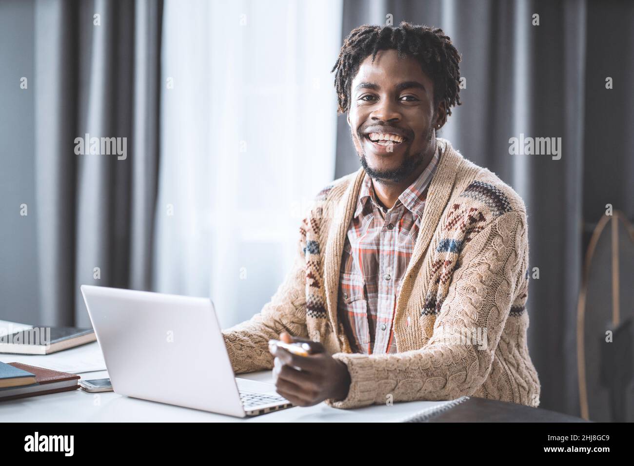 Uomo africano che lavora da casa utilizzando un computer portatile seduto accanto alla finestra. Giovane imprenditore che lavora sul suo portatile a casa. Studio di studenti afroamericani a casa durante il blocco. Foto Stock
