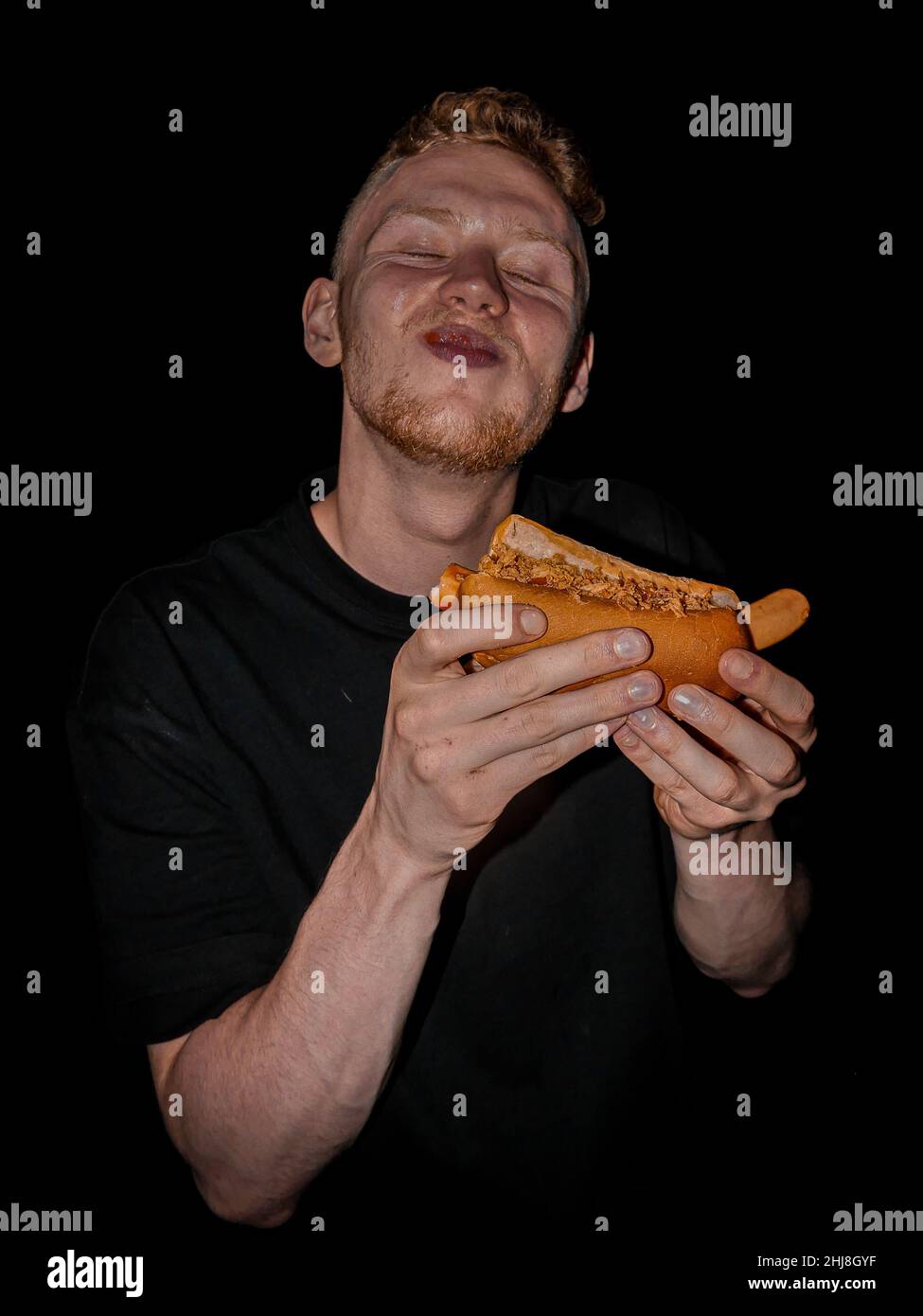 Giovane uomo che mangia il suo hotdog davanti ad uno sfondo scuro Foto Stock