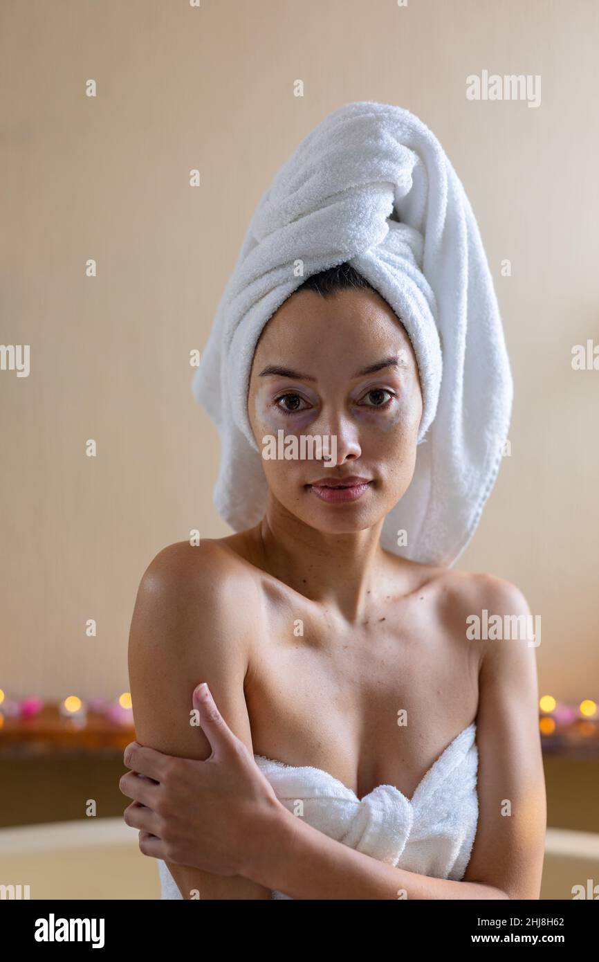 Ritratto di giovane donna biraciale con asciugamano avvolto sulla testa e il corpo seduta contro la parete in spa. spa, cura del corpo, sensualità, stile di vita, vitiligo e. Foto Stock