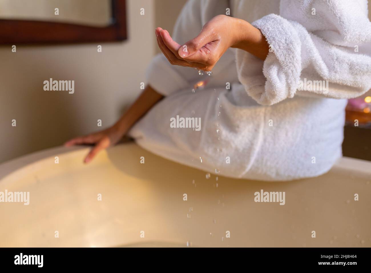 Giovane donna biraciale che indossa l'accappatoio toccando l'acqua mentre si siede nella vasca idromassaggio. centro benessere, cura del corpo, stile di vita e benessere. Foto Stock