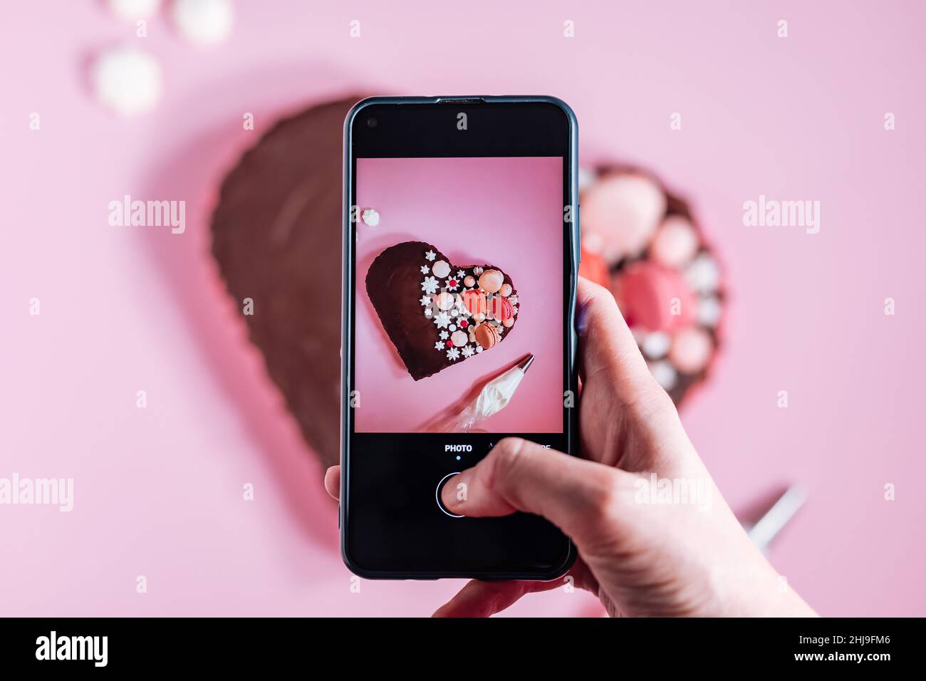 Fotografando la torta a forma di cuore rosa con glassa al cioccolato, meringhe e amaretti in cima come decorazione. Posizionato su sfondo rosa. San Valentino. Foto Stock