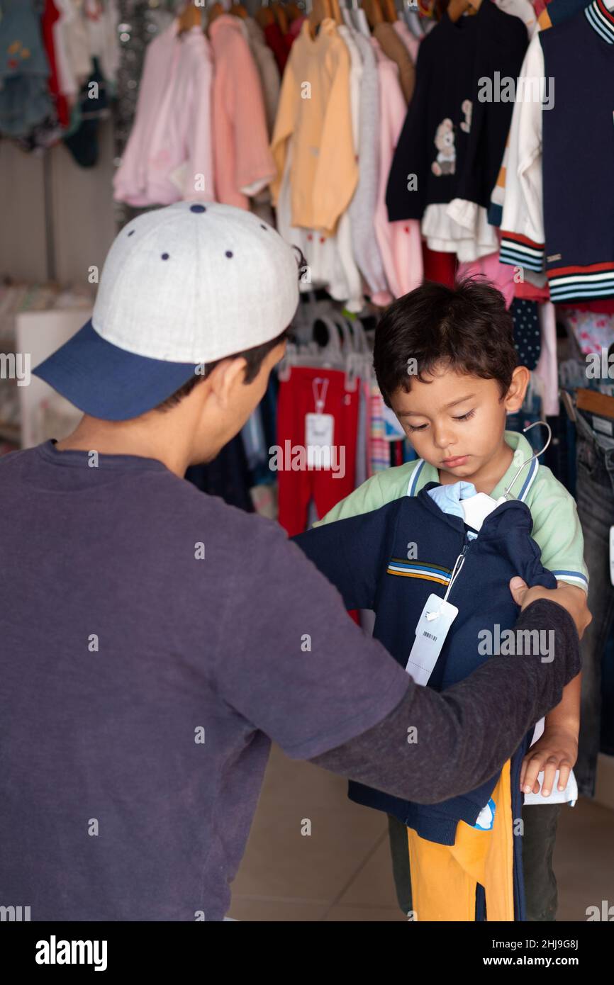 Foto verticale di un giovane adulto che acquista abiti per suo figlio. Uomo latino che taglia una camicia o un maglione per suo figlio in un negozio di abbigliamento. Misurare gli indumenti sopra Foto Stock