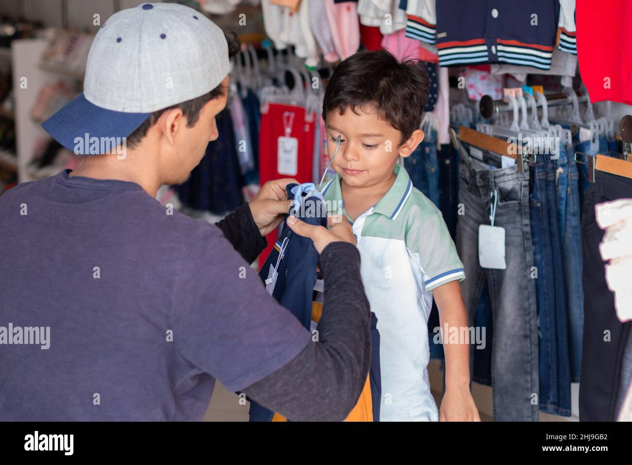 Giovane adulto che acquista abiti per suo figlio, uomo latino che taglia una camicia o un maglione per suo figlio in un negozio di abbigliamento. Foto Stock