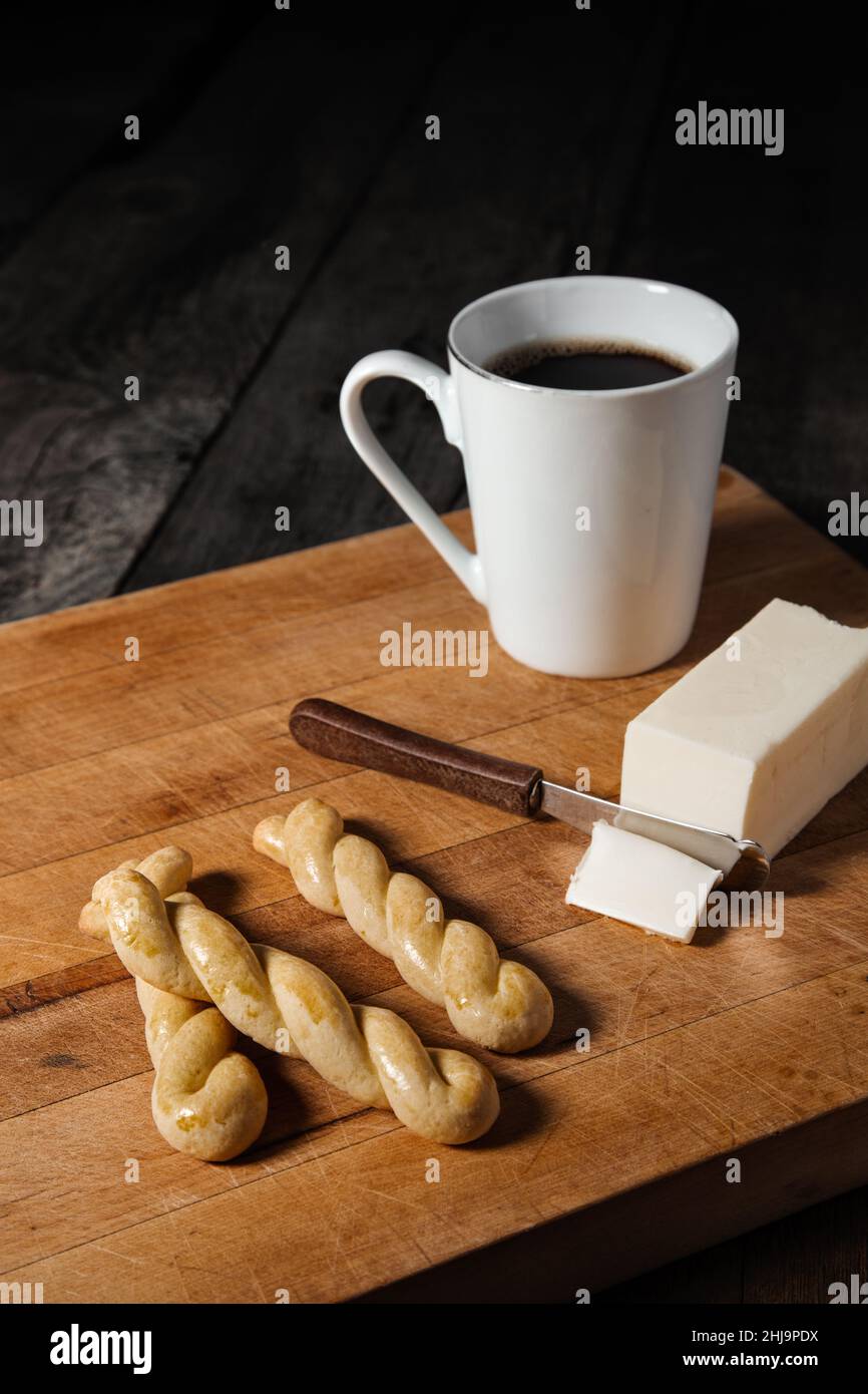 Una colazione a base di bastoncini di pane al forno, un bastone di burro e una tazza di caffè scuro posto su un tavolo di legno nero. Foto Stock