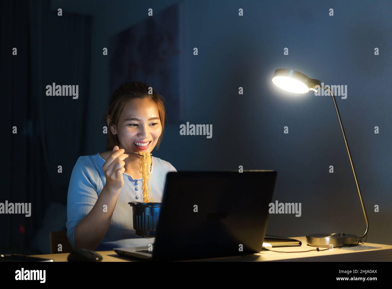 Donna d'affari intelligente freelance asiatica che mangia spaghetti istantanei mentre lavora su un computer portatile nel soggiorno a casa di notte ragazza asiatica felice seduta sopra Foto Stock