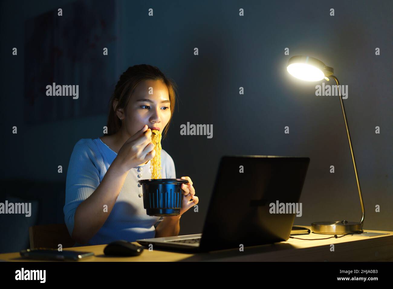 Donna d'affari intelligente freelance asiatica che mangia spaghetti istantanei mentre lavora su un computer portatile nel soggiorno a casa di notte ragazza asiatica felice seduta sopra Foto Stock