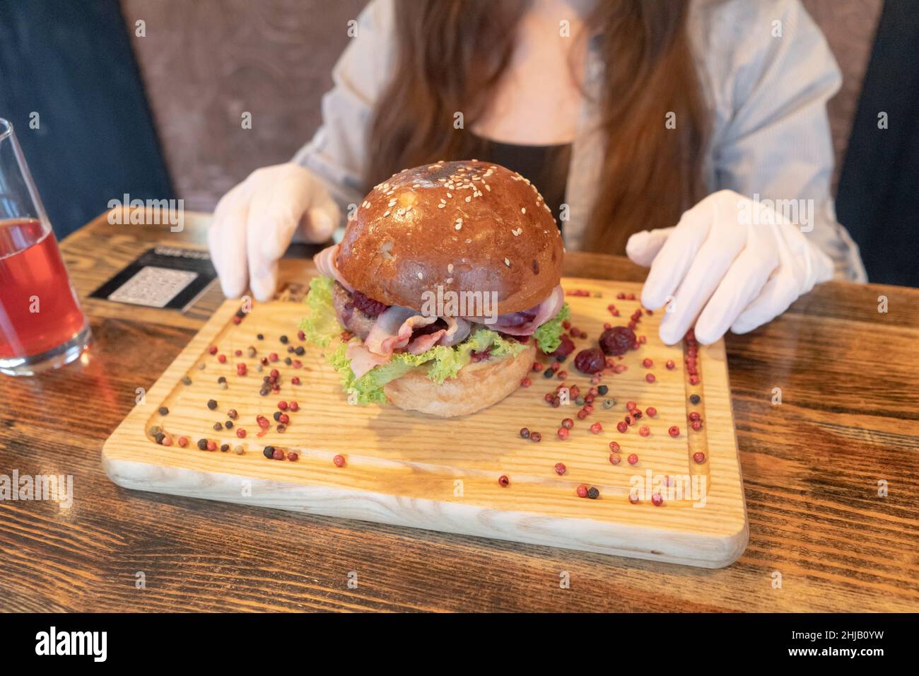 Grande hamburger bello su un bordo di legno con piselli di pepe. Una ragazza si siede ad un tavolo che indossa guanti di gomma prima di mangiare. Fast food. Foto Stock