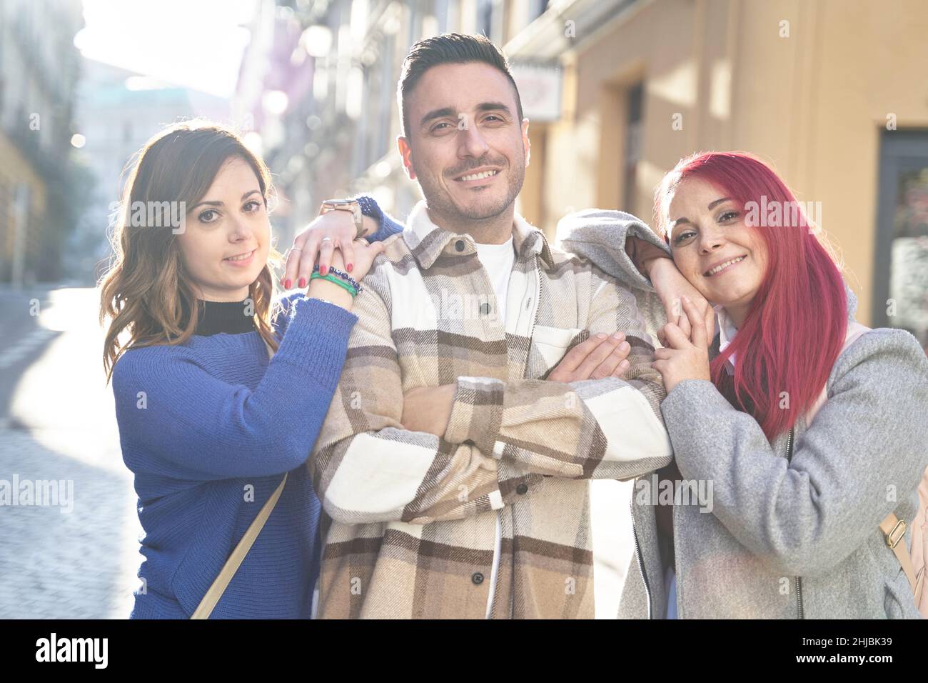 Donne positive appoggiate sulle spalle dell'uomo con le braccia incrociate e guardate la macchina fotografica con il sorriso mentre si trova in città in giorno di sole Foto Stock